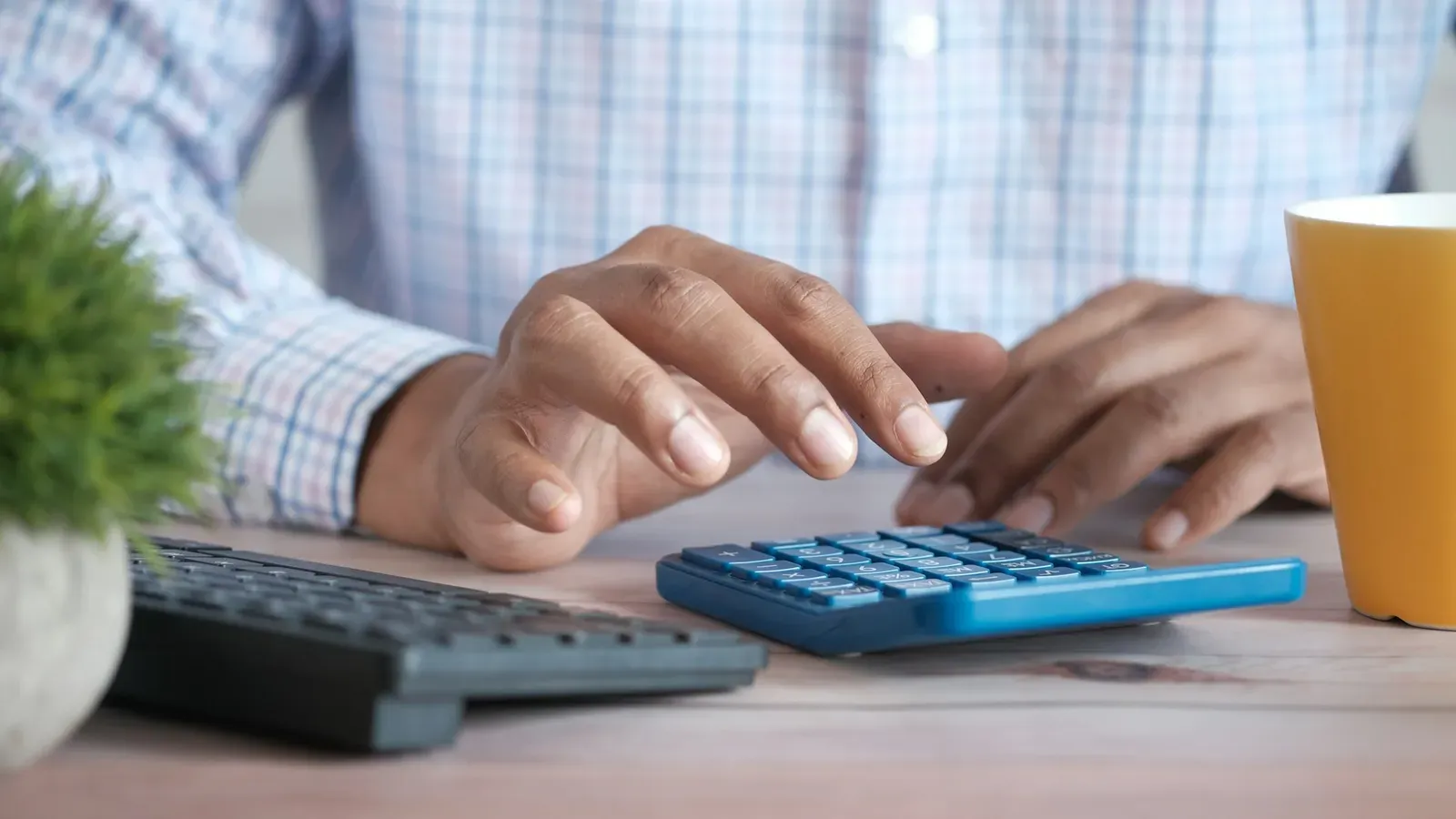 Mãos digitando numa calculadora azul sobre uma mesa de madeira, ao lado de um teclado, de um vaso de planta e de uma caneca amarela