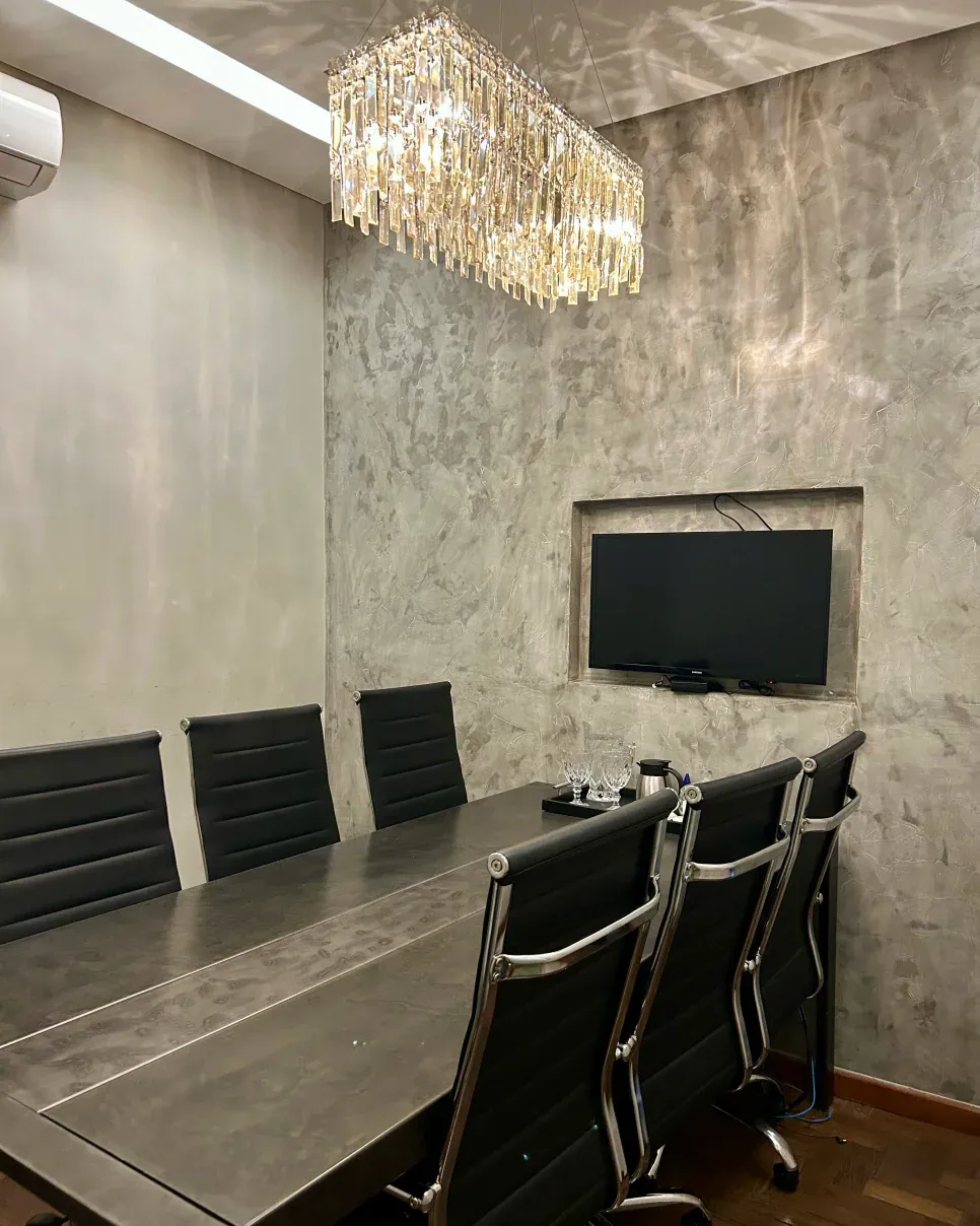 Sala de reunião com mesa longa escura, cadeiras pretas, televisão embutida na parede e lustre de cristal